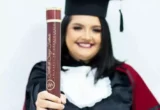 Estudante com traje de formatura posa para o retrato com o diploma na mão.