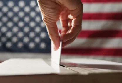 Detalhe de mão depositando voto em urna com bandeira dos Estados Unidos ao fundo.