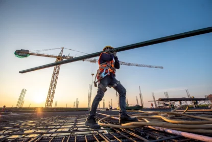 Trabalhador da construção civil carrega viga em meio a obra de prédio.