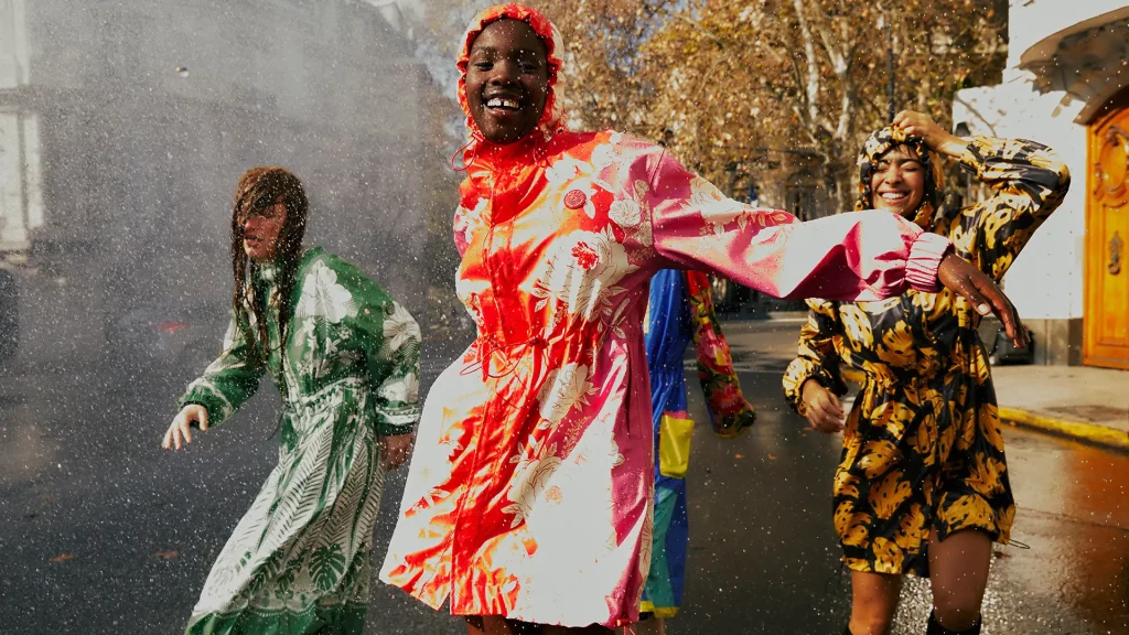 Grupo de pessoas em ambiente urbano sob garoa, vestindo roupas coloridas, destacando capa de chuva floral vermelha e rosa.