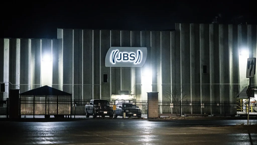 Fachada noturna do edifício industrial JBS com iluminação intensa e portão de segurança na entrada.