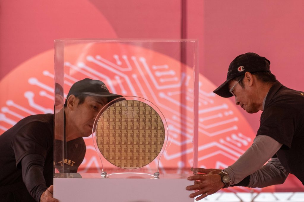 Dois homens manuseiam expositor transparente com um wafer de silício. Fundo vermelho com padrão de circuito.