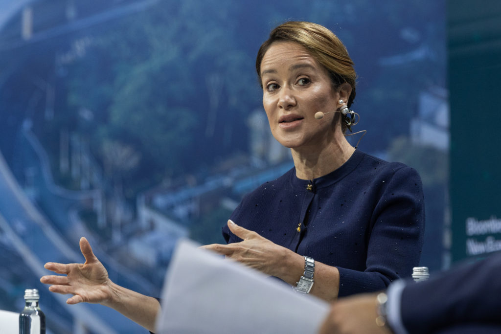 Mulher de cabelo castanho e blusa escura fala e gesticula. Fundo desfocado com paisagem verde-azulada.