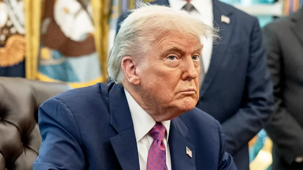 Donald Trump, de terno azul com pin da bandeira, sentado em um escritório. Ele tem um olhar sério e pensativo.