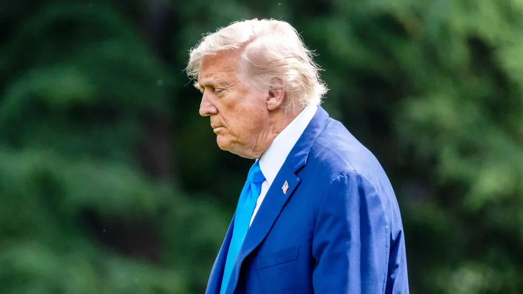 Donald Trump em terno azul e gravata, em perfil, olhando para baixo. Fundo borrado verde.
