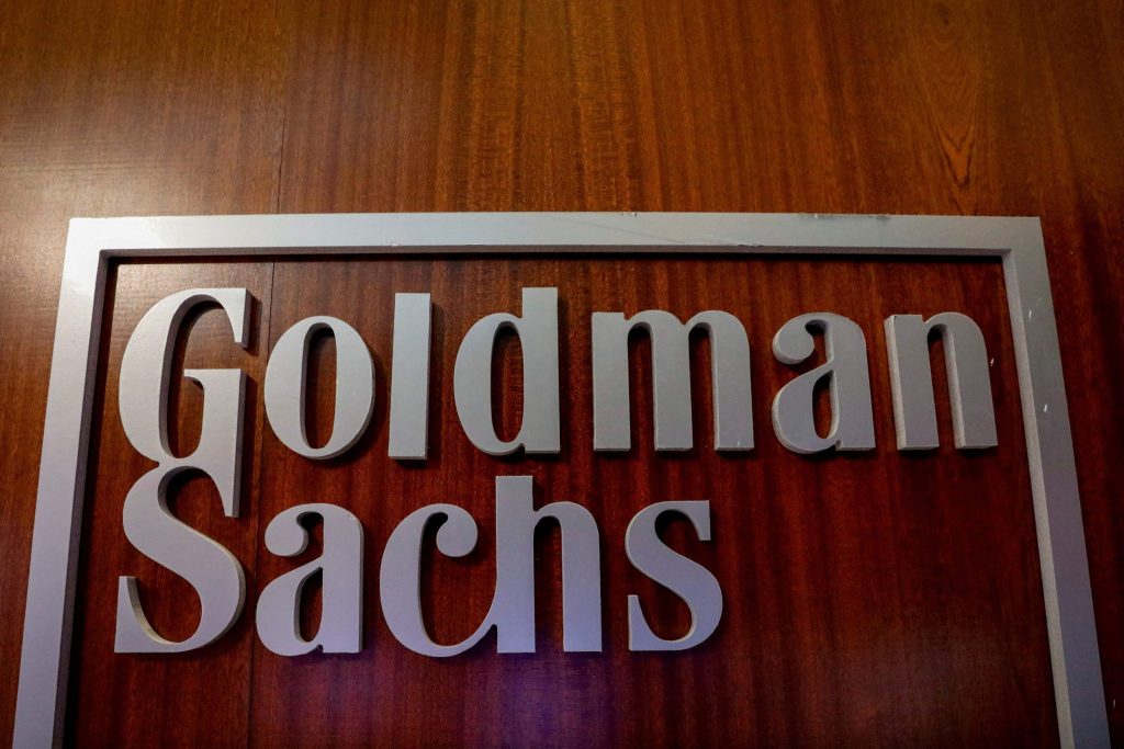 Letreiro "Goldman Sachs" com letras cinzas e moldura sobre fundo de madeira escura.