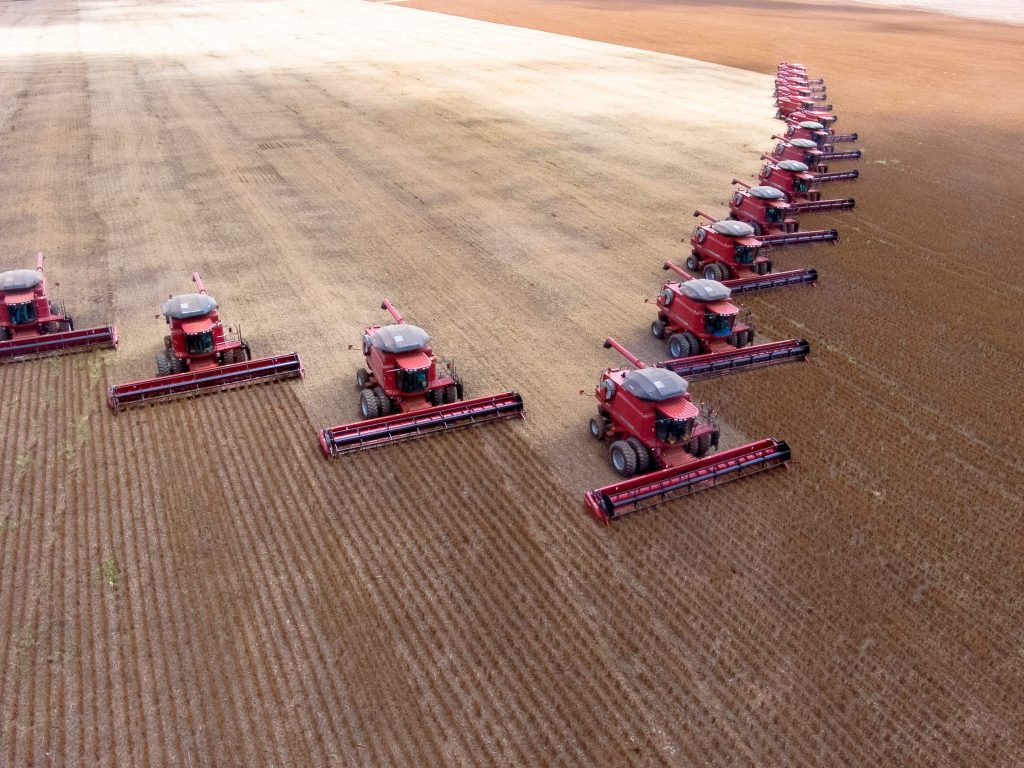 Vista aérea de várias colheitadeiras vermelhas alinhadas, trabalhando na colheita de um campo agrícola extenso.