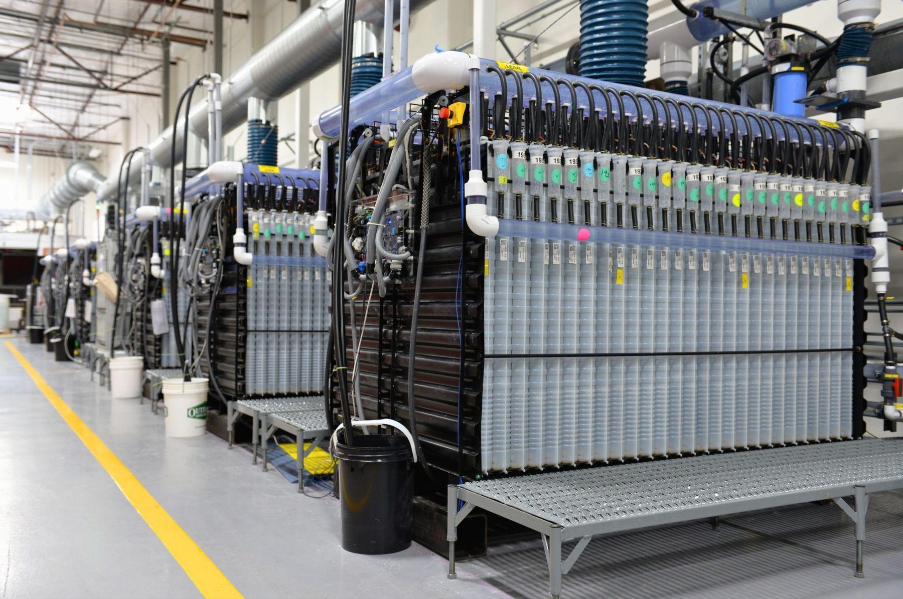 Interior de uma instalação industrial com fileiras de módulos de bateria de ferro-ar conectados por tubos, cabos e sistemas de circulação de fluidos