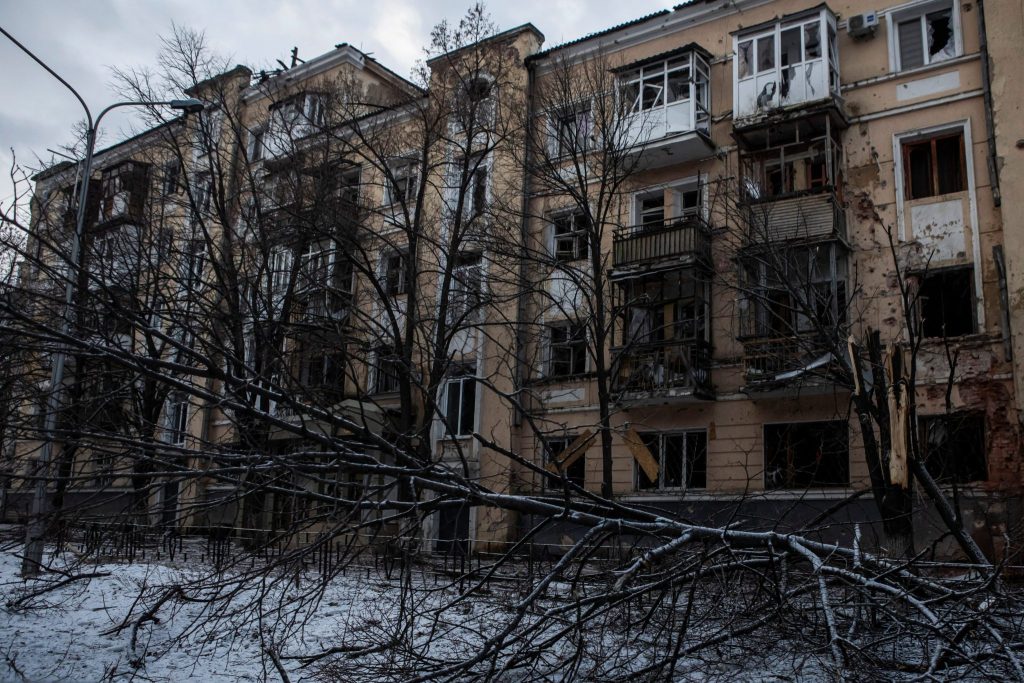 Prédio residencial destruído com janelas estilhaçadas e varandas danificadas, em paisagem de inverno com neve e galhos.