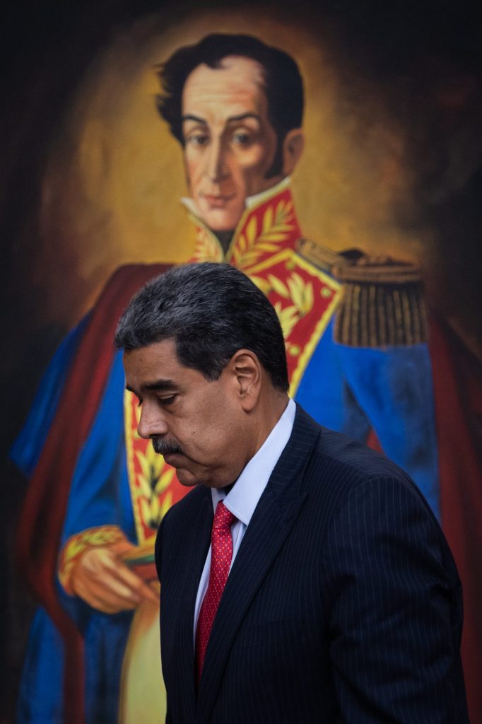 Nicolás Maduro de terno escuro e gravata vermelha caminha, com pintura de Simón Bolívar ao fundo.