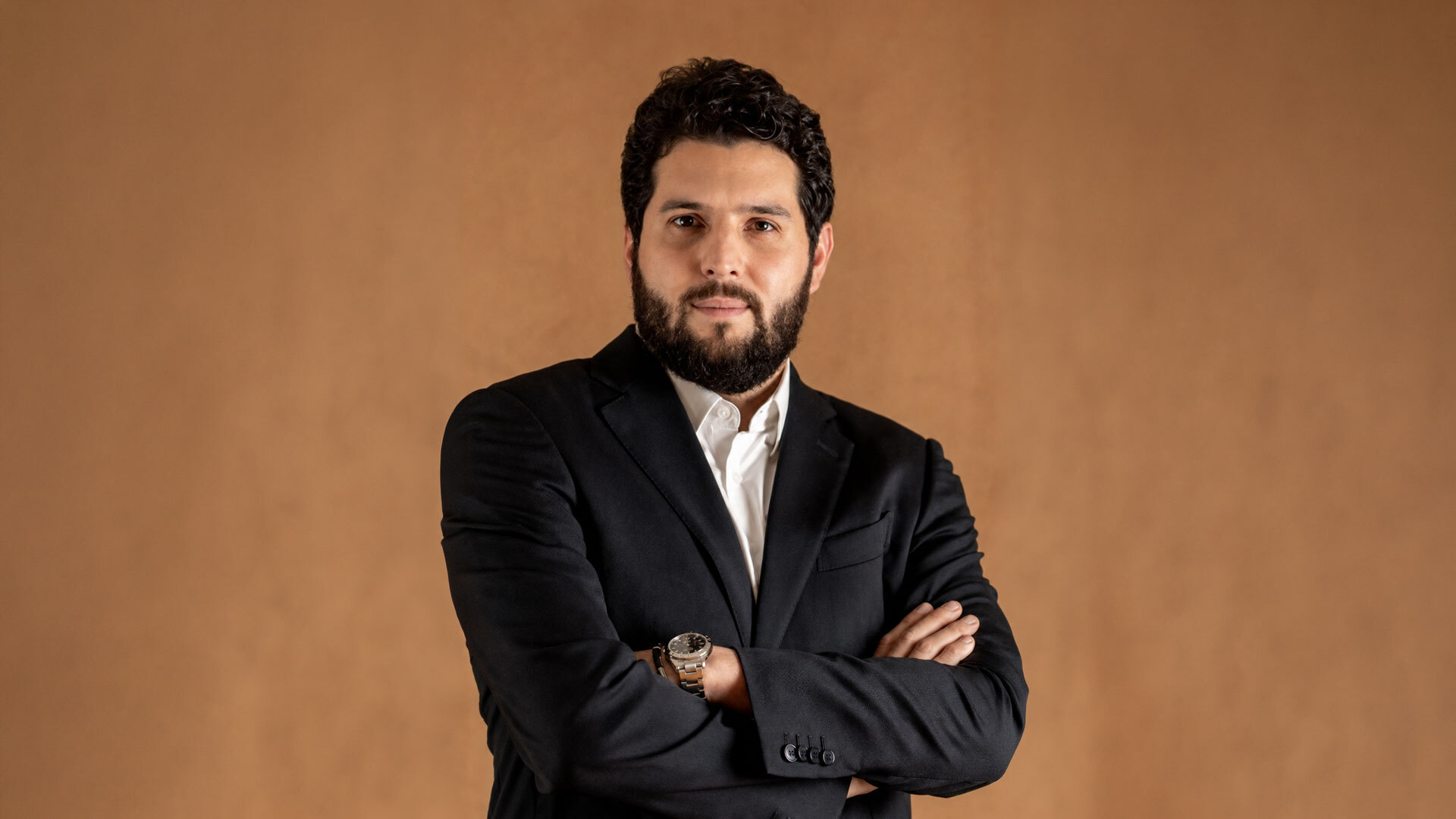 Retrato de Aguinaldo Filho usando terno escuro e camisa branca, com os braços cruzados, olhando para a câmera diante de um fundo marrom.