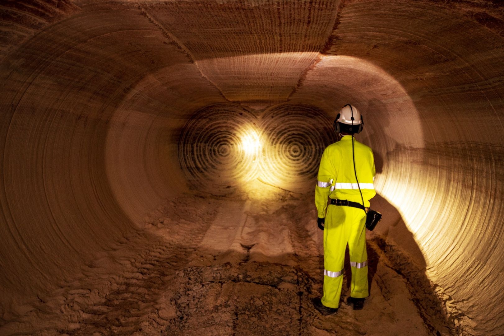 Trabalhador com uniforme de alta visibilidade e capacete observa um túnel subterrâneo da mina de potássio de Taquari-Vassouras, em Sergipe, iluminado por uma lanterna e com marcas circulares de perfuração nas paredes de rocha
