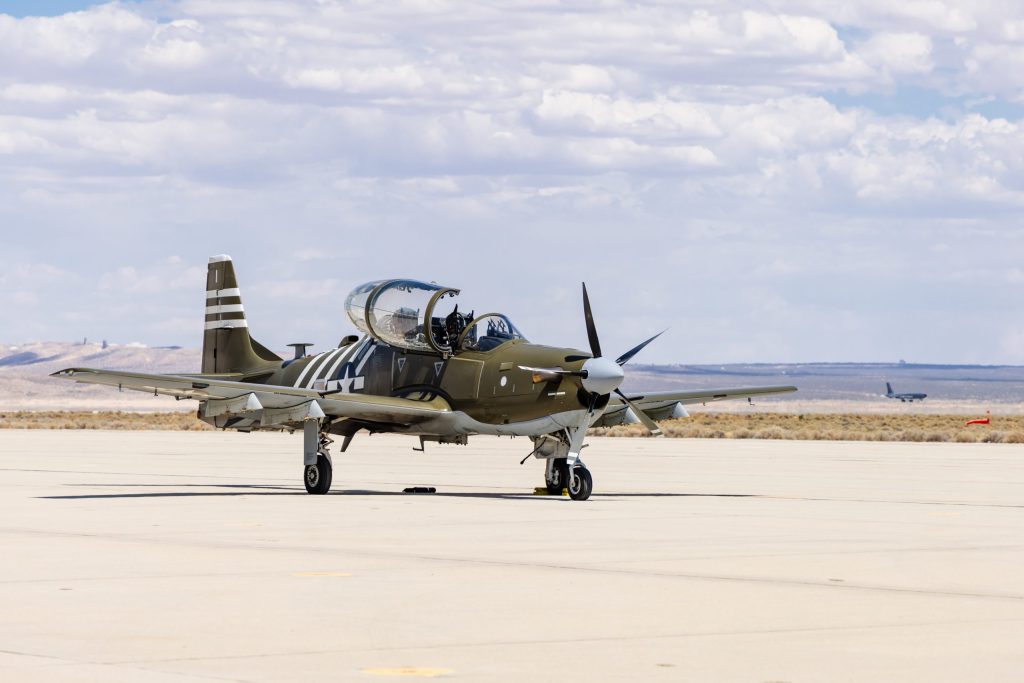 Avião militar verde-oliva com hélice, parado em pátio de aeroporto.